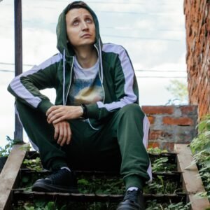 A man in a green tracksuit sits contemplatively on leafy steps against a brick wall, looking upward outdoors.