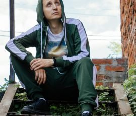 A man in a green tracksuit sits contemplatively on leafy steps against a brick wall, looking upward outdoors.